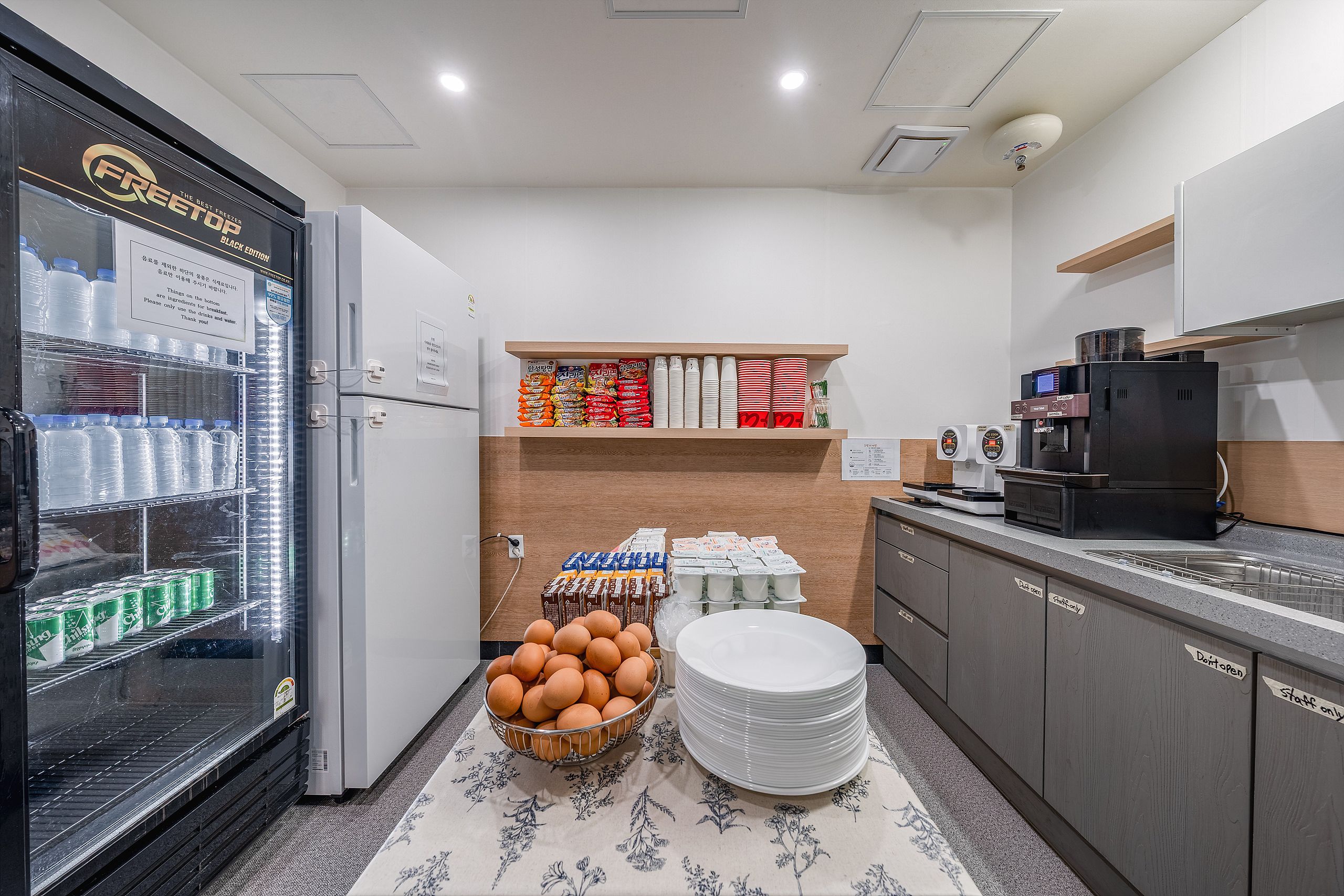 A kitchen area with a refrigerator, shelves with packaged goods, a coffee machine, and a sink.