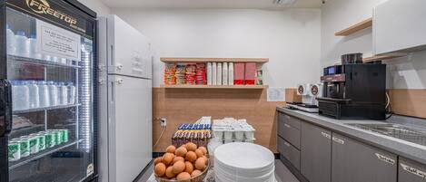 A kitchen area with a refrigerator, shelves with packaged goods, a coffee machine, and a sink.
