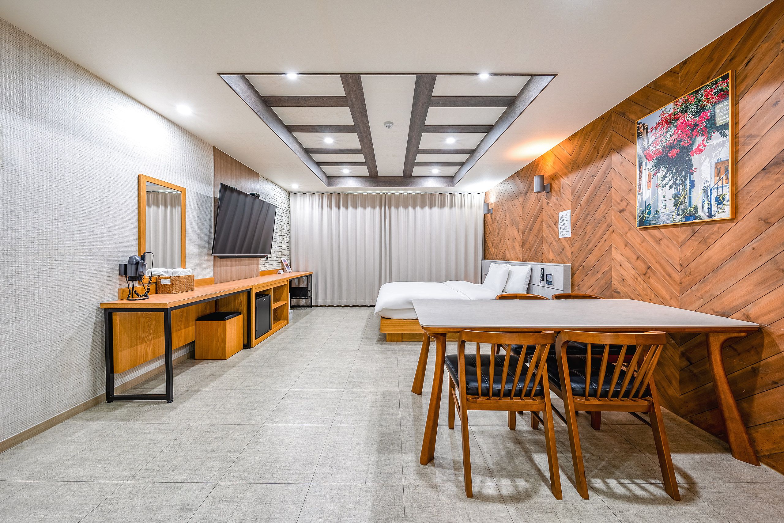 A modern hotel room with a wooden herringbone wall, a large bed, a desk with a flat-screen TV, and a dining area with wooden chairs.