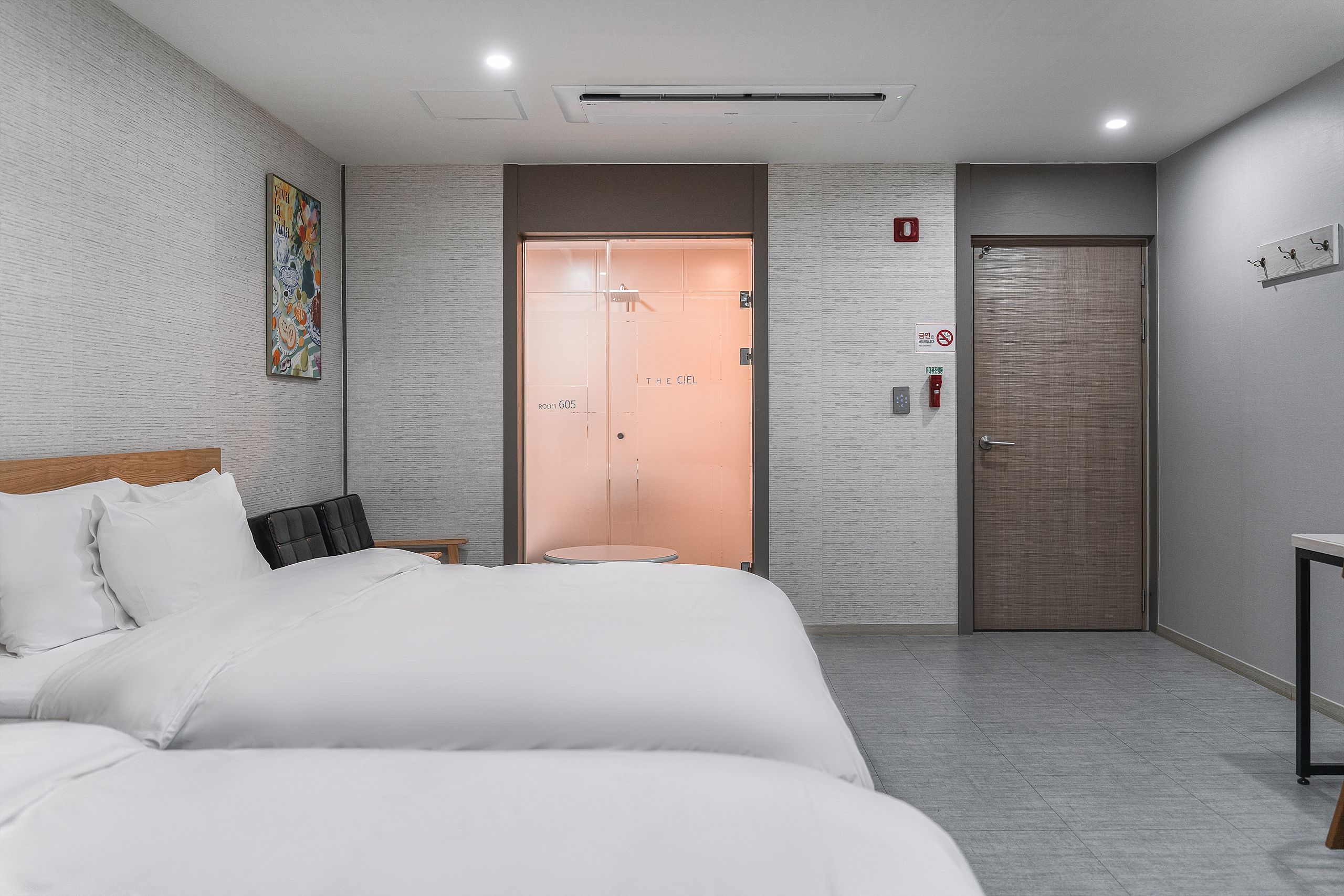 A hotel room with a bed, a small table, a wall-mounted artwork, a door, and a wall-mounted fire alarm.