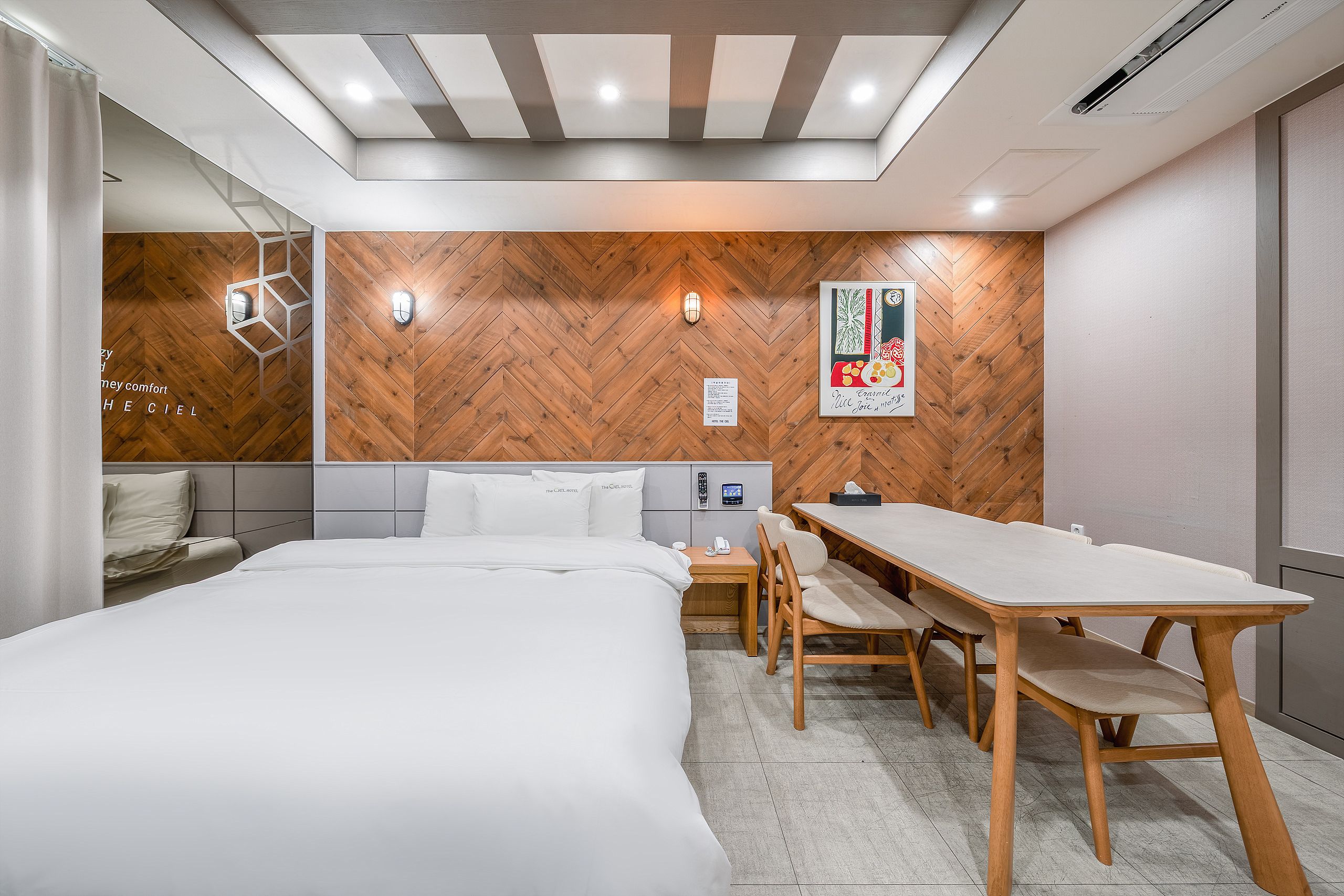 A hotel room with a bed, a desk, and a chair, a wooden herringbone wall, and a ceiling with recessed lighting.