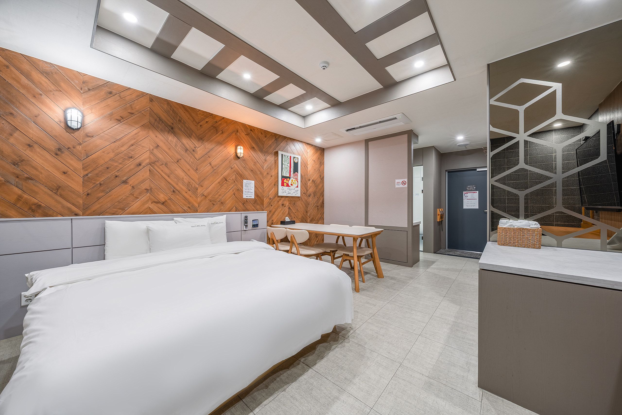 A hotel room with a large bed, a wooden accent wall, a small table with chairs, and a bathroom with a geometric glass partition.