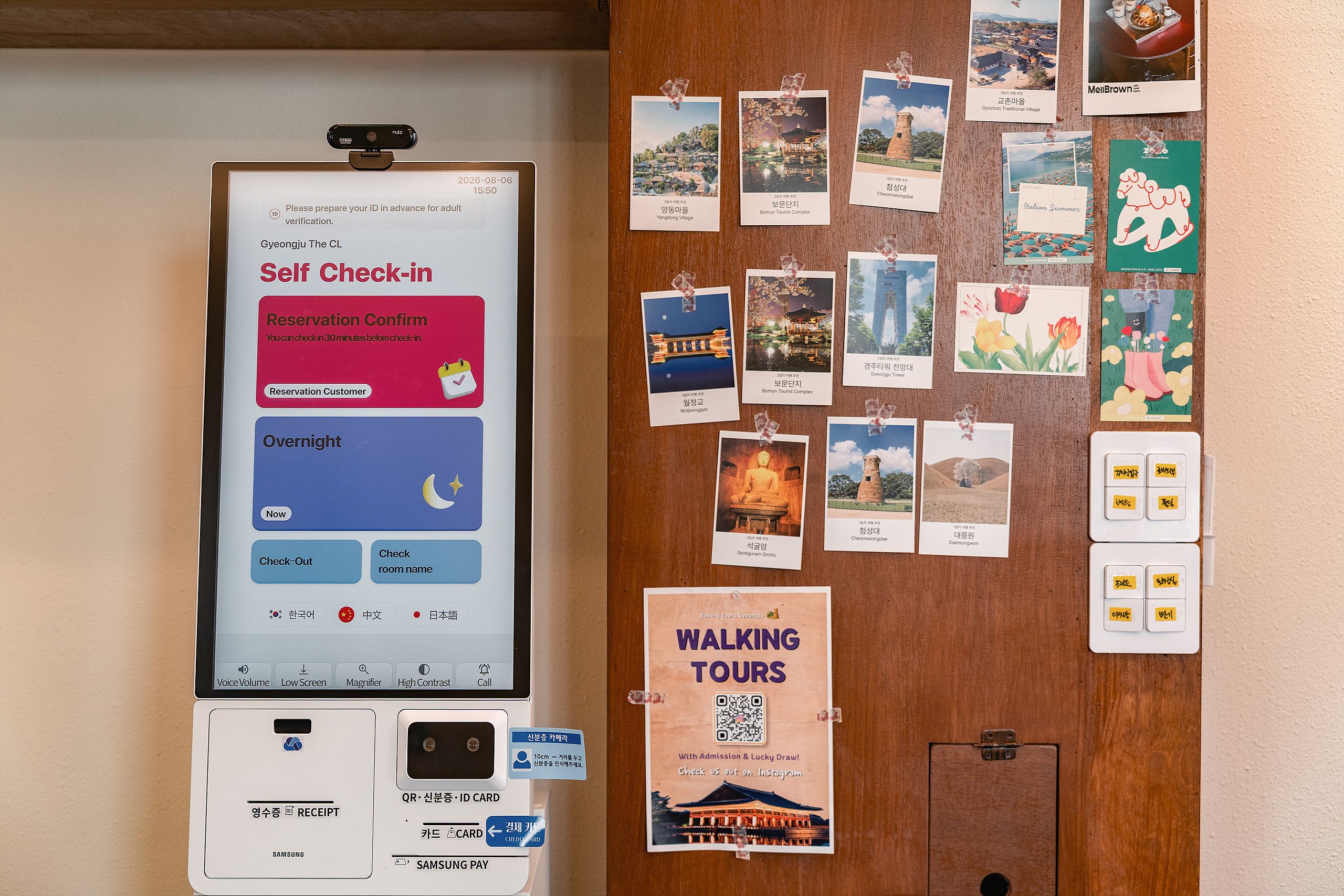 Un quiosco de auto check-in con una pantalla, un terminal de pago Samsung y una pared con varios carteles de viajes.