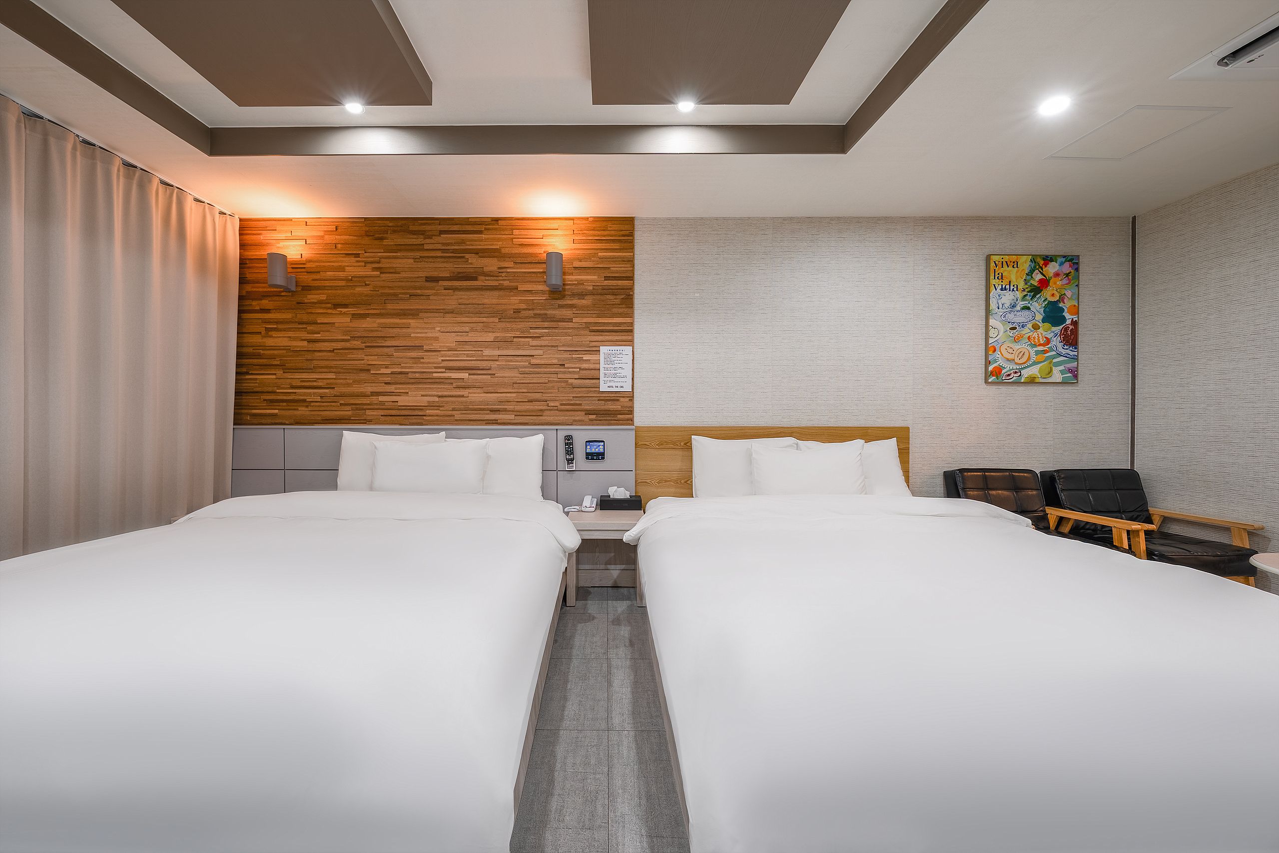 A hotel room with two beds, a wooden accent wall, a painting, and a desk with a chair.