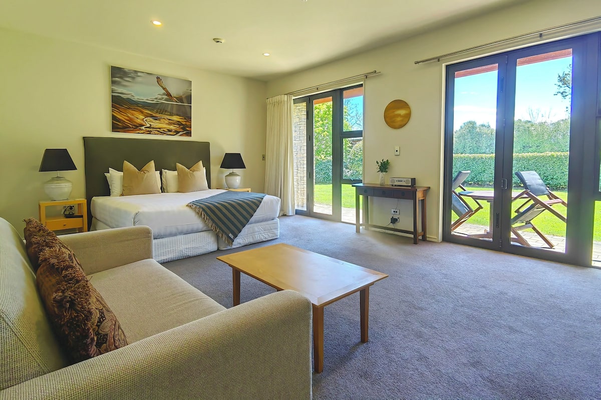 Habitación de hotel con una cama grande, un sofá, una mesa de centro y un escritorio con una silla. Hay una ventana grande con vistas a un jardín.