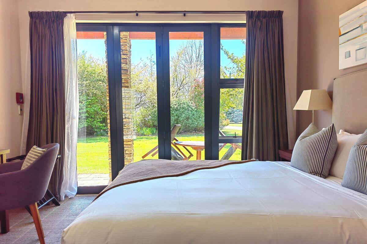 Habitación de hotel con una cama grande, una silla, una mesa y vistas a un jardín a través de amplios ventanales.