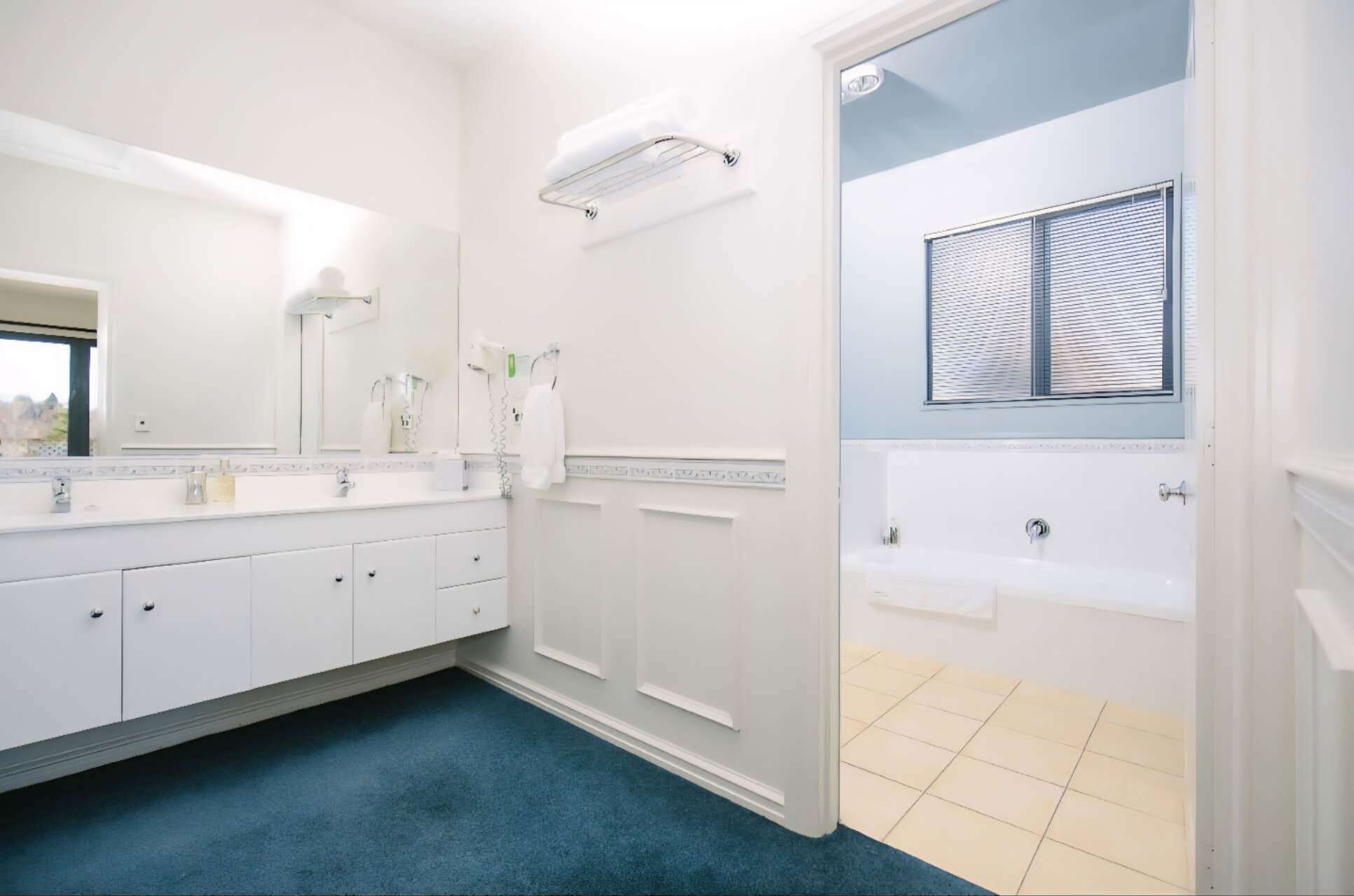 A bathroom with a white vanity, a large mirror, a towel rack, and a window with blinds.