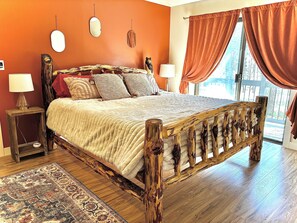 Un lit en bois avec une tête de lit, deux tables de chevet avec des lampes, un tapis à motifs et une vue sur les arbres par la fenêtre.