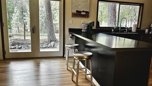 Une cuisine moderne dotée d’un grand îlot central, de tabourets de bar et offrant une vue sur une zone boisée grâce à des portes vitrées.