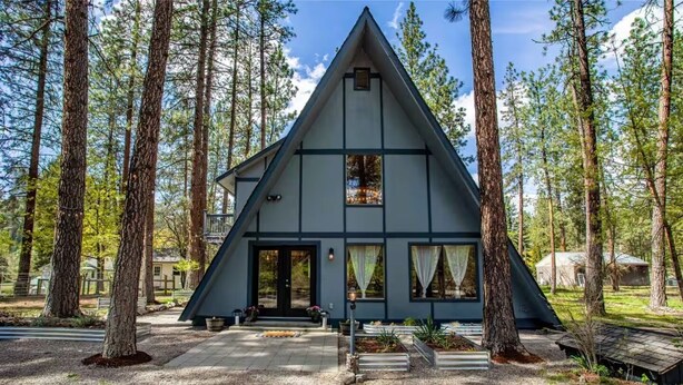 Une cabane triangulaire dotée d’une véranda, entourée de grands arbres.