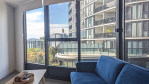 Una sala de estar moderna con un sofá azul, una pequeña mesa de madera y ventanas amplias que ofrecen vistas a un paisaje urbano y zonas verdes.