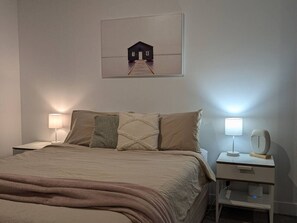 A bedroom with a bed, two bedside tables, a nightstand, a wall-mounted lamp, and a framed picture of a house on a pier.