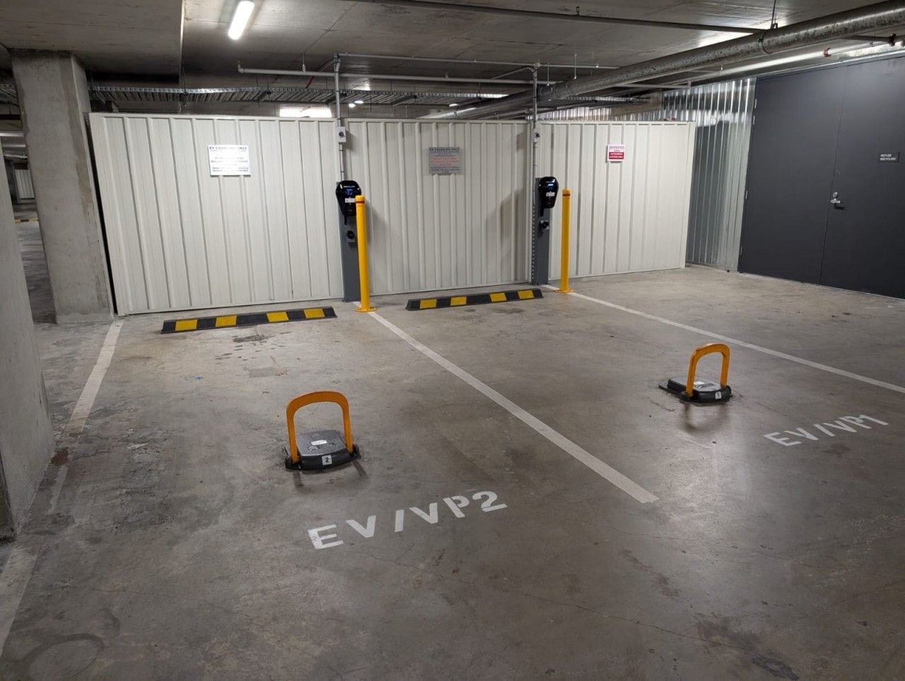 A parking garage with two parking spaces marked EV/VP2, equipped with yellow bollards and a barrier gate.