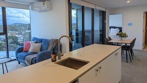 Une cuisine moderne dotée d’un grand îlot central, un coin repas avec une table et des chaises, un canapé avec des coussins et une vue sur la ville grâce à de grandes fenêtres.
