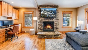 A cozy living room with a stone fireplace, wooden flooring, and a large window overlooking a snowy landscape. - Moose Run Lodge – Slope-Side Luxury Retreat at Whitefish Mountain (Whitefish)