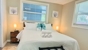 A neatly made bed with white linens, a pillow with a floral design, and rolled towels on the bed. There is a wooden bench at the foot of the bed, and two bedside lamps.