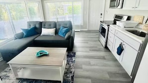 A modern living room with a sectional sofa, a coffee table, and a kitchen area with white cabinets and appliances.