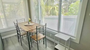 A dining area with a wooden table and chairs, a vase with flowers, and large windows with blinds.