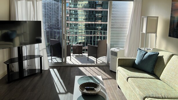 Uma sala de estar moderna com televisão de tela plana, sofá, mesa de centro e uma varanda com vista para um edifício alto.