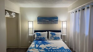 Quarto de hotel com cama e espalha cama com tema havaiano, pintura de uma cena tropical acima da cama, e mesa de cabeceira branca com abajur.