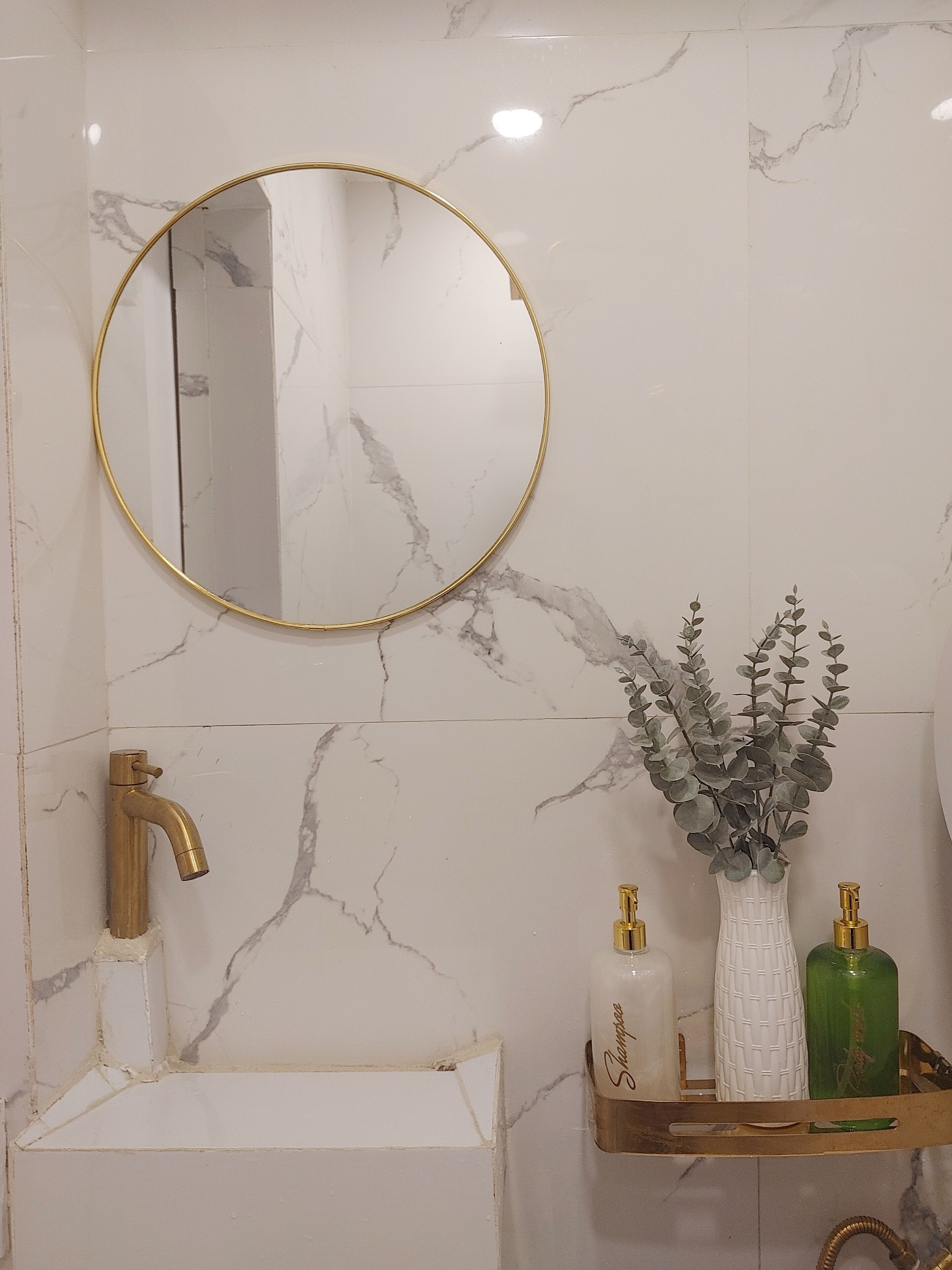 A bathroom with a marble wall, a gold-framed mirror, a gold faucet, and a shelf with toiletries.