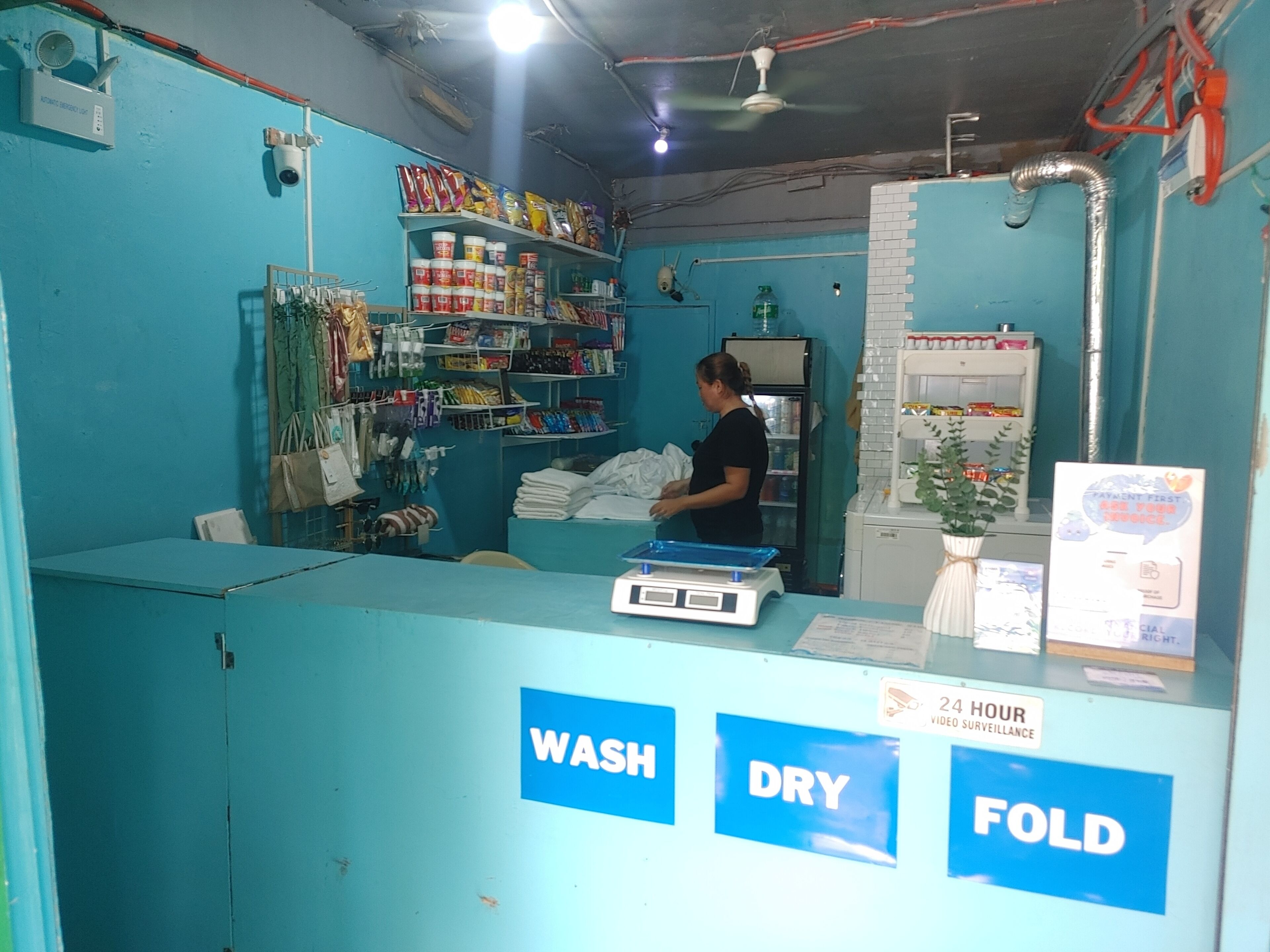 A small, brightly lit laundry facility with blue walls and a counter labeled with wash, dry, and fold services.