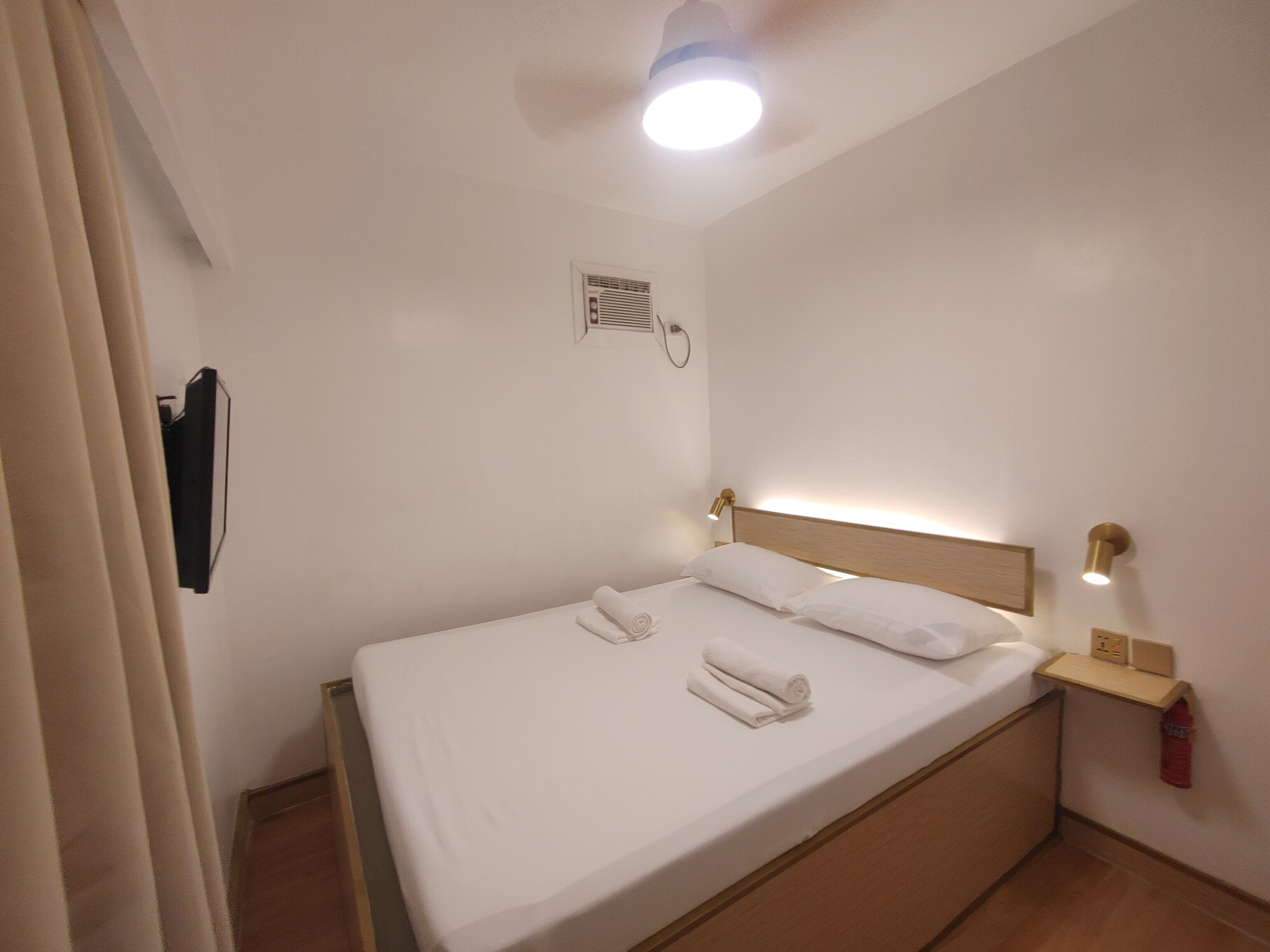 A neatly made bed with white linens and two rolled towels, a bedside table with a lamp, and a ceiling light.