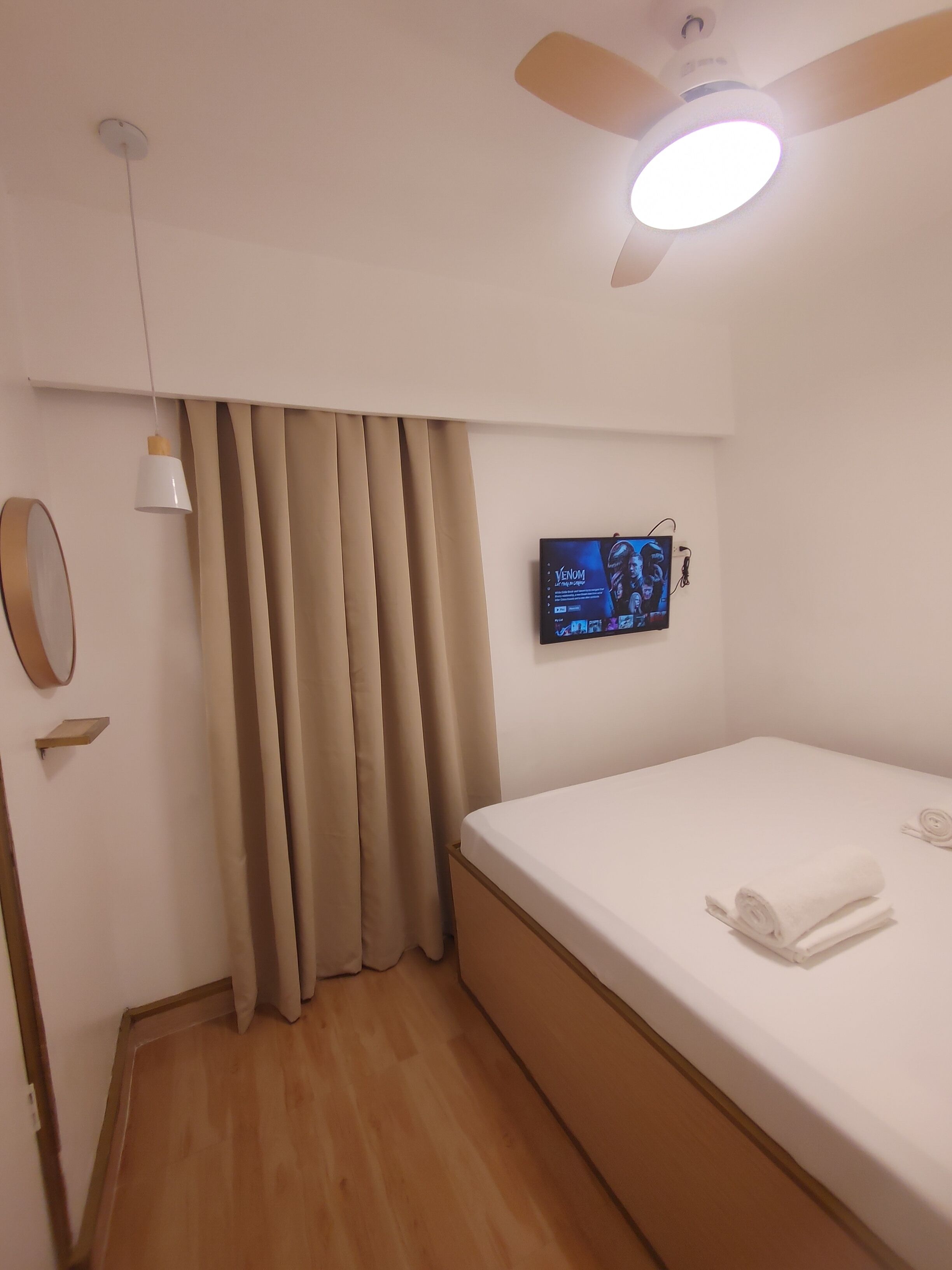 A hotel room with a bed, a ceiling fan, a TV mounted on the wall, a mirror, and a window with curtains.