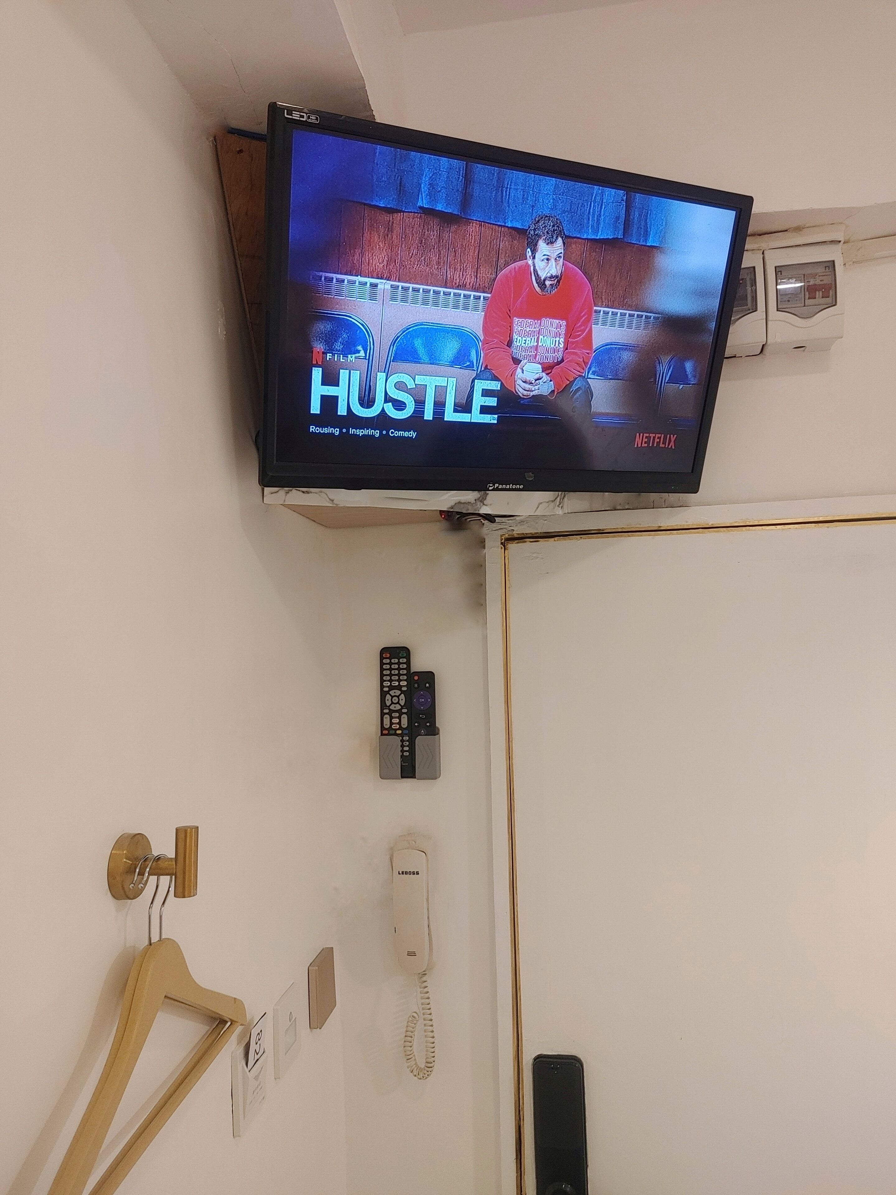 A television mounted on a wall displaying the 'Hustle' show, with a remote control, a telephone, and a hanger on the wall below.
