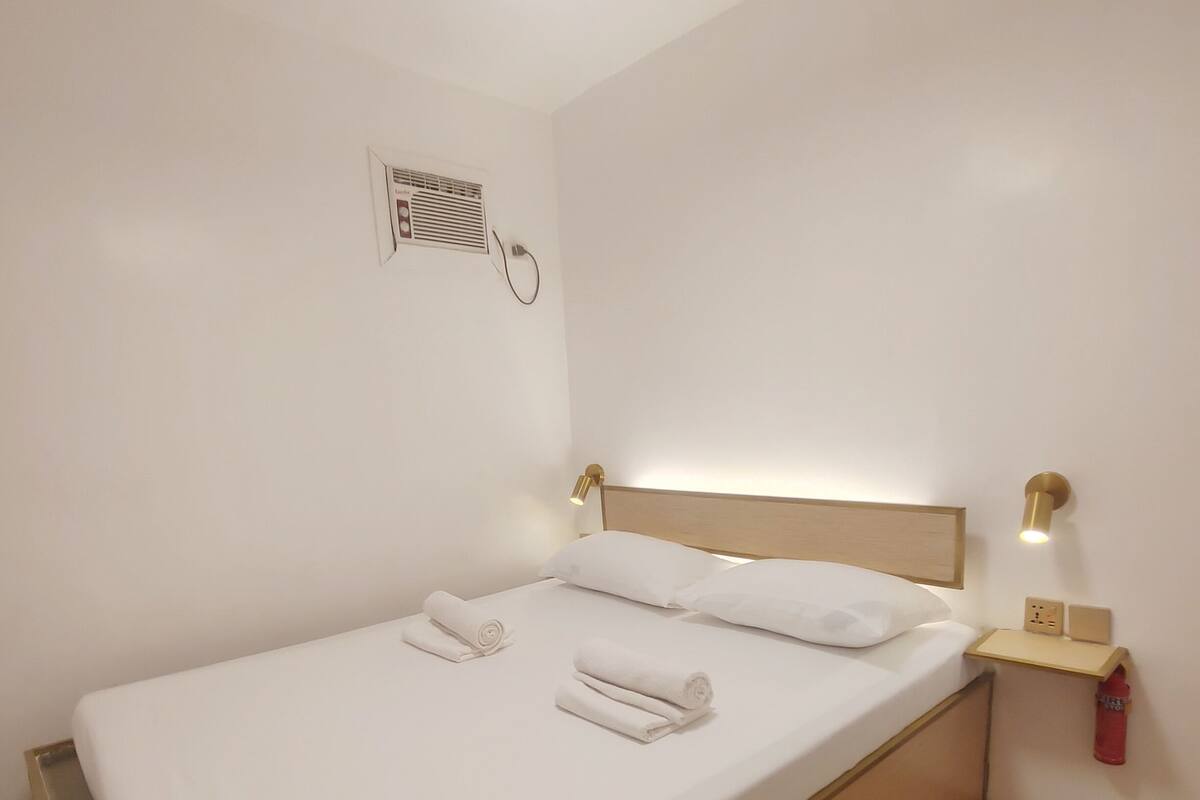 Ein ordentlich bezogenes Bett mit weißen Leinentüchern und aufgerollten Handtüchern, ein Deckenventilator, ein Nachttisch mit Lampe sowie eine Wand-Klimaanlage.