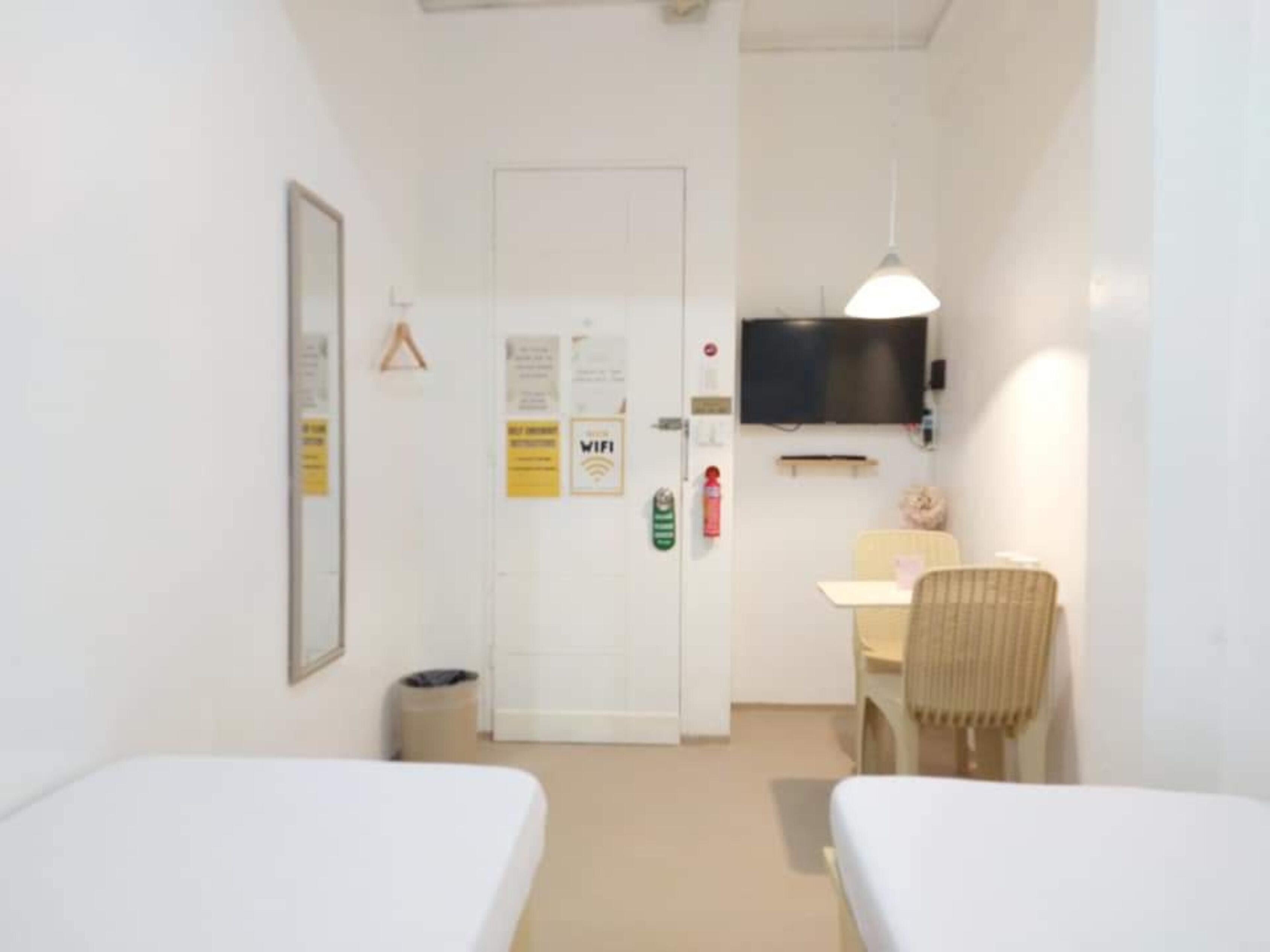 A small, clean room with two single beds, a desk with a chair, a TV mounted on the wall, and a door with a sign.