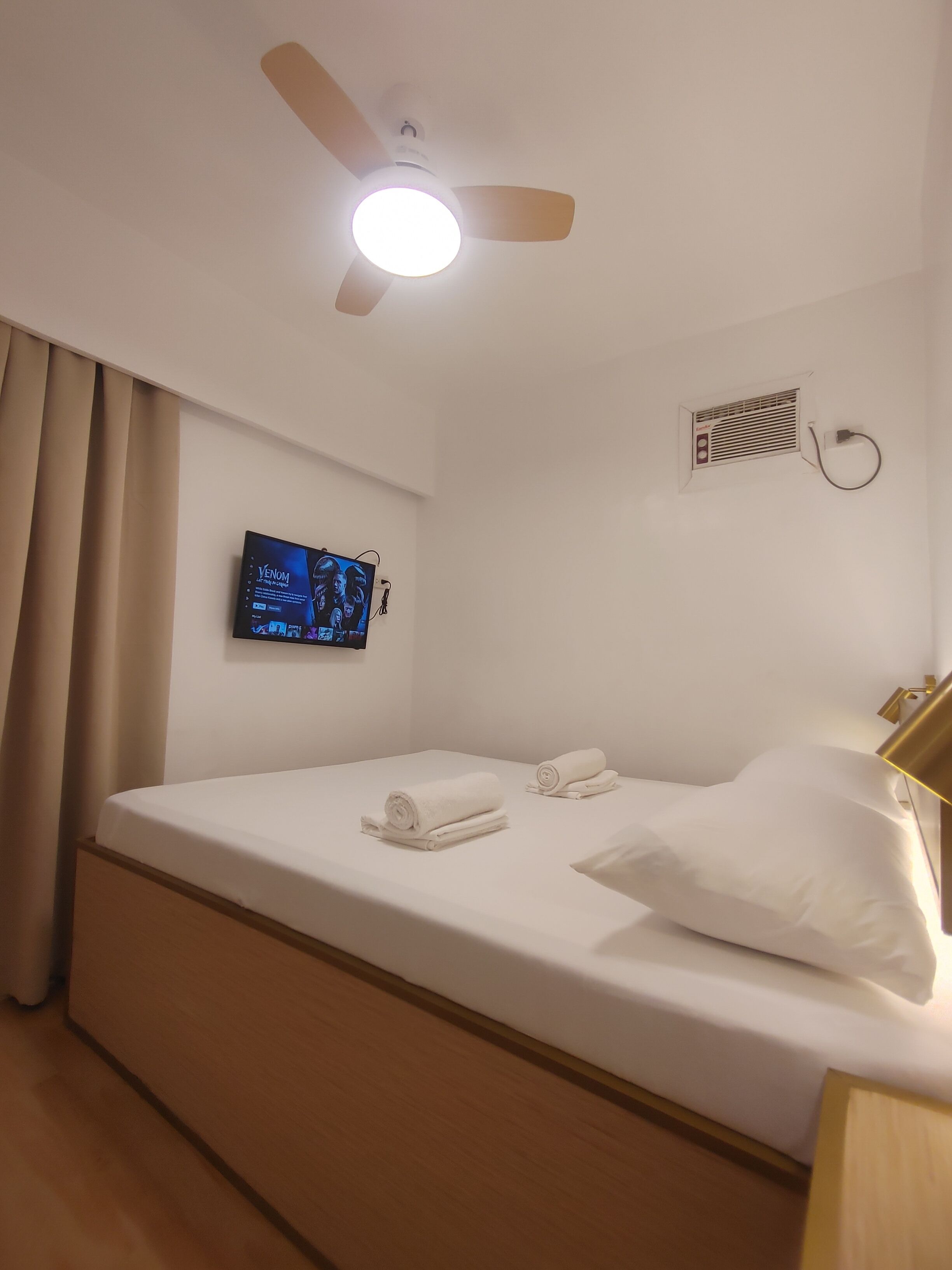 Una habitación de hotel con cama, ventilador de techo, televisor montado en la pared y aire acondicionado de pared.