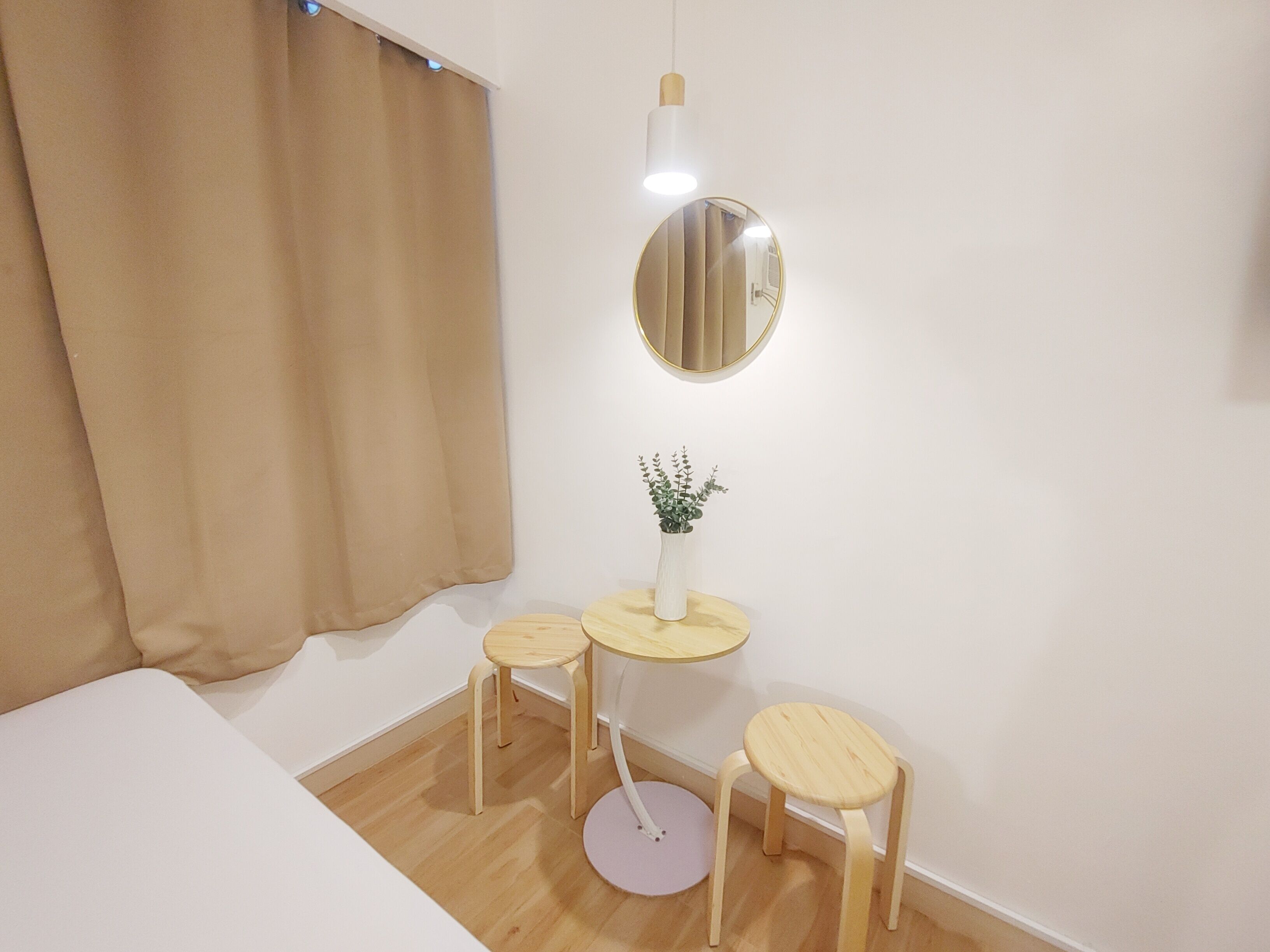 Una habitación pequeña y moderna con una cama, dos taburetes, una mesita con una planta y un espejo redondo en la pared.