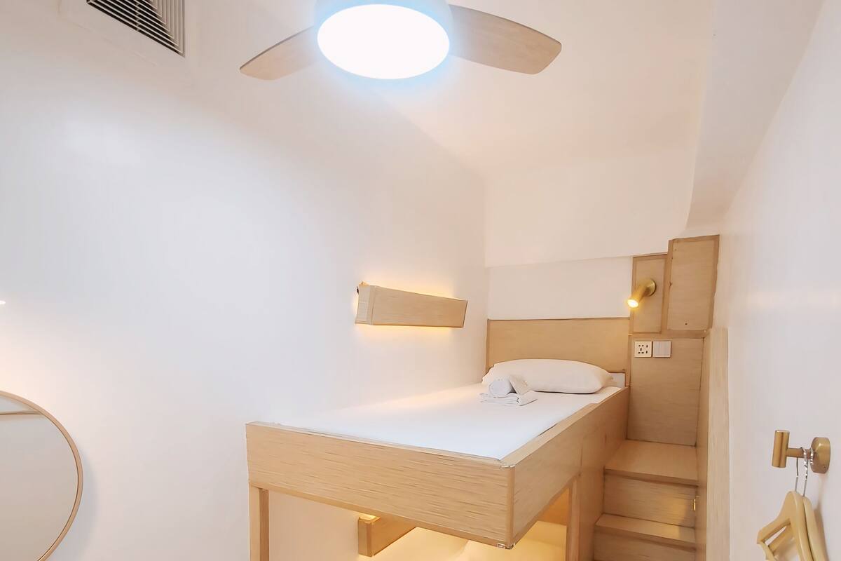 Ein kleines Zimmer mit einem Etagenbett, einem Schreibtisch mit zwei Stühlen, einem kleinen Tisch, einem Deckenventilator und einer Treppe.