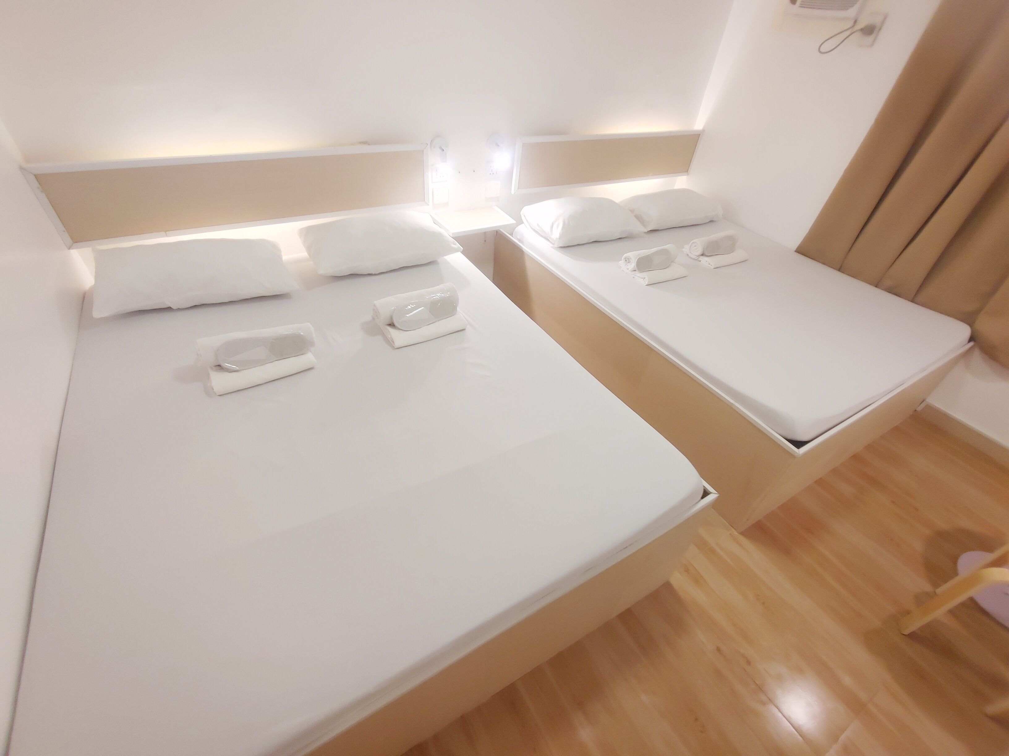Dos camas individuales con sábanas y almohadas blancas, suelo de madera y una mesita de noche con lámpara.