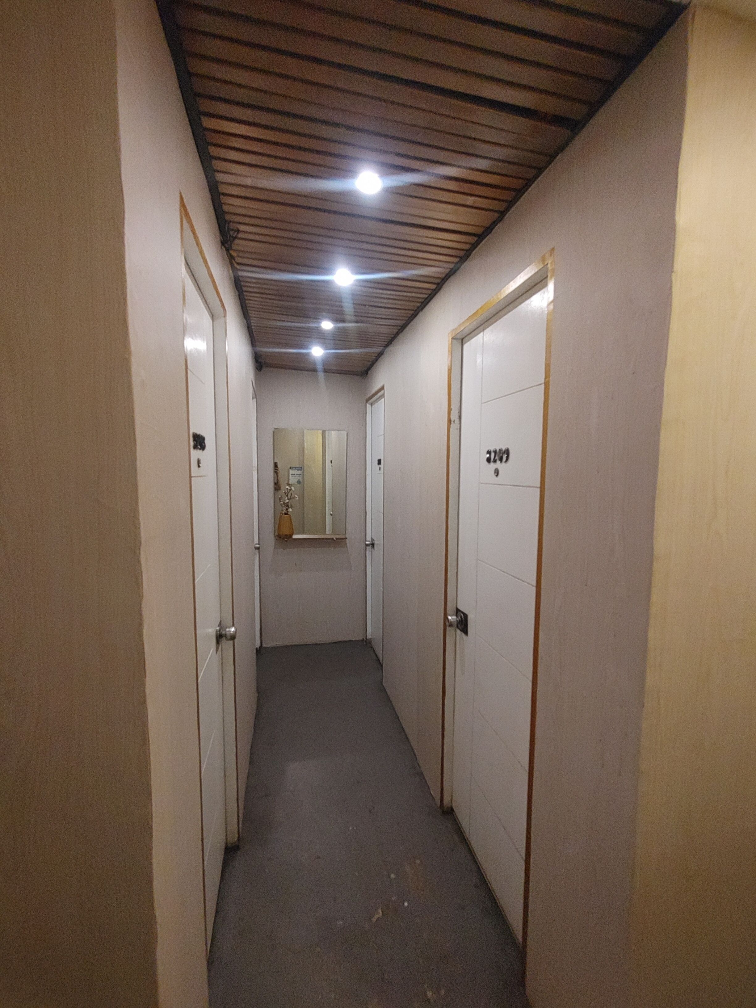 A narrow hallway with two doors on each side, a mirror on the left wall, and a ceiling with wooden beams and recessed lighting.