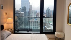 Een slaapkamer met een groot raam met uitzicht over de stad, een bed, een bedlamp en een klein krukje.