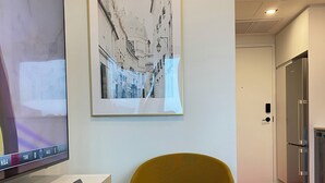 Een modern interieur met een flatscreen televisie op een witte standaard, een mosterdgele stoel, een stadsgezicht in een lijst en een rood tapijt dat leidt naar een keuken met roestvrijstalen apparatuur.