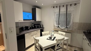 A modern kitchen with white cabinets, a dining table with chairs, a TV mounted above the counter, and a window with curtains. - Cozy 2 X room, kitchen bathroom apartment in the Karl Marx Siedlung in Worms 1st floor (Worms)