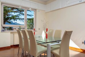 A dining area with a glass table and beige chairs, large windows with a view of trees and buildings, and a radiator on the wall. - Pine House (Sorrento)
