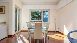 A modern dining area with a glass table and chairs, large windows with a view of trees, and a wooden floor. - Pine House (Sorrento)