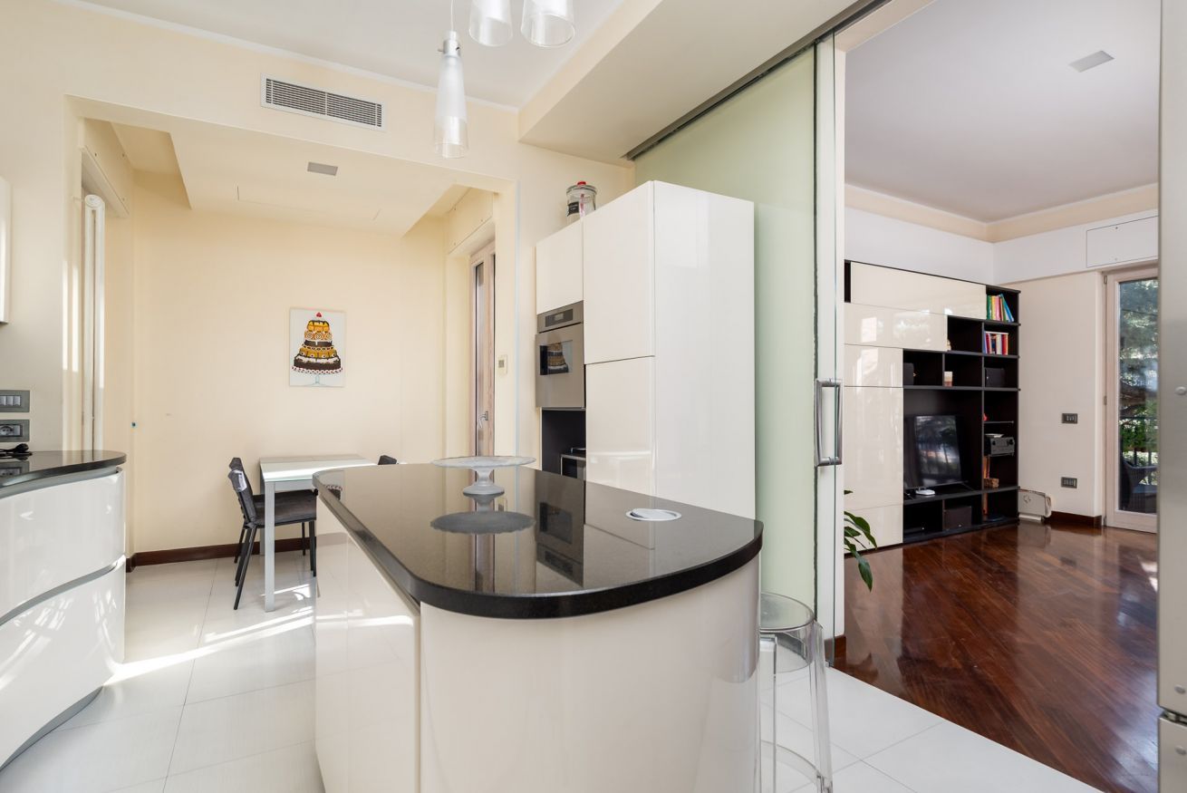 A modern kitchen with a central island, a dining area with a table and chairs, and a living area with a TV and bookshelves.