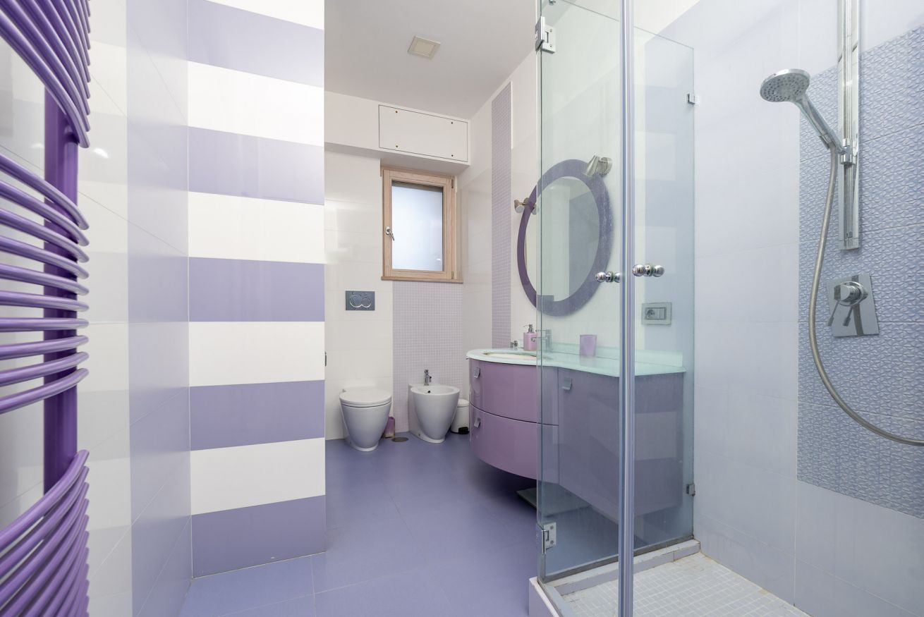 A modern bathroom with a glass shower enclosure, a round mirror, and a purple-striped wall.