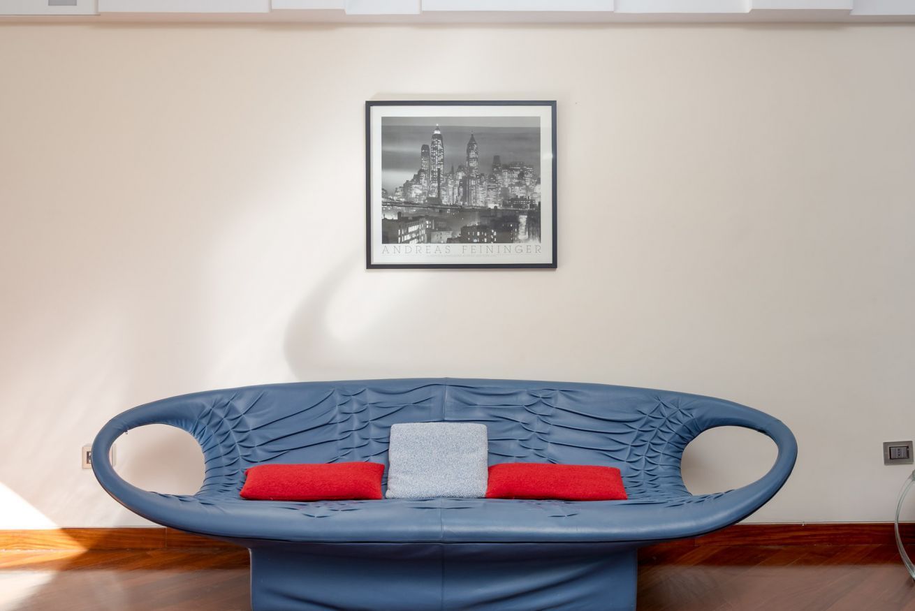 Una habitación con un sofá azul, un cuadro con una vista urbana enmarcada y una almohada roja.