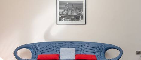Una habitación con un sofá azul, un cuadro con una vista urbana enmarcada y una almohada roja.