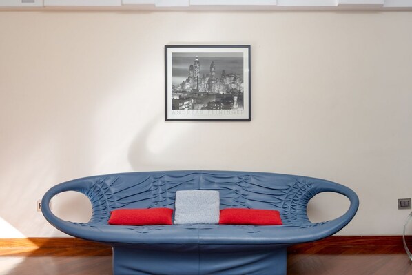 Una habitación con un sofá azul, un cuadro con una vista urbana enmarcada y una almohada roja.