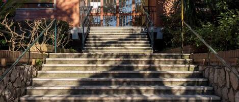Un conjunto de escalones de piedra que conducen a la entrada de un edificio, con barandilla de metal y vistas al verde circundante.