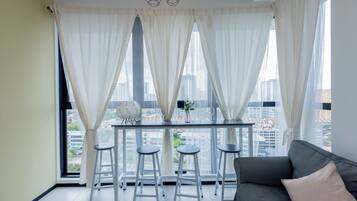 A modern room with a dining table, two bar stools, a sofa, and a city view through large windows with sheer curtains.