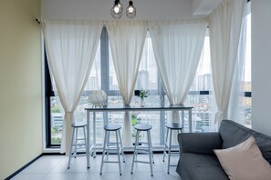 A modern room with a dining table, two bar stools, a sofa, and a city view through large windows with sheer curtains. - Urban Suite Penang (George Town)