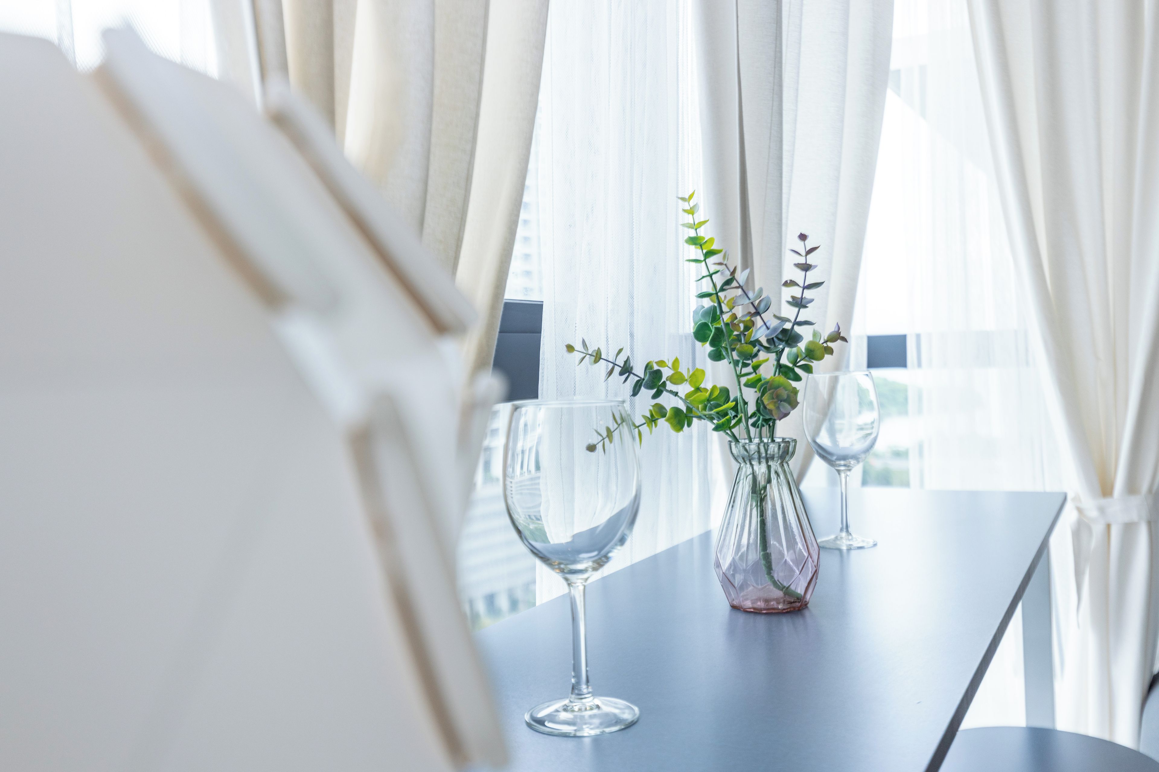 Een tafel met twee glazen wijnglazen en een vaas met groene bladwerk, voor een achtergrond van transparante witte gordijnen en een raam met uitzicht op gebouwen.